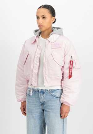 Vrouw met een lichtroze bomberjack met meerdere zakken en een rode "Remove Before Flight" label op de mouw, daaronder een grijze hoodiesweater.