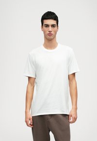 Vit kortärmad t-shirt i bomull med rundhalsdesign, visad på en modell mot en enkel ljus bakgrund.