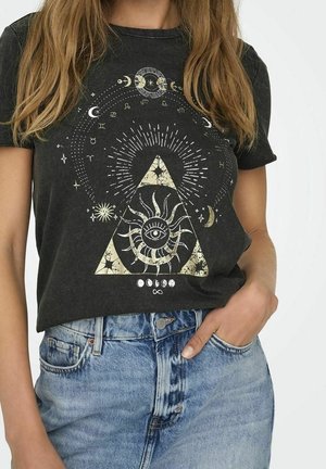 Vrouw draagt een donker T-shirt met een mystiek ontwerp van zon, maan, ster en sterrenbeeld symbolen, gecombineerd met blauwe spijkerbroek.