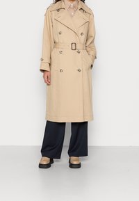 Beige trench coat with double-breasted design, large buttons, and a belt. Paired with dark wide-leg pants and tan shoes with black soles.
