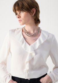 Hvid bluse med flæset halsudskæring, lange ærmer og et glat, let stof. Modellerer sølv lagdelte kæd necklaces og mørke bukser.