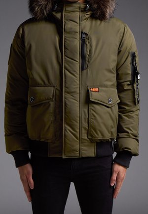 Mand iført olivengrøn polstret jakke med pelsbetonet hætte, flere lynlåse og klaplommer samt sorte ribstrikkede manchetter og kant.