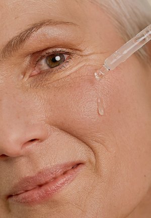 Clear facial serum being applied with a pipette to a cheek. Skin is light-toned with visible texture and fine lines. Soft lip color.