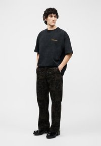 Mann mit lockigem Haar, der ein schwarzes übergroßes T-Shirt, dunkel gemusterte Hose mit Blumenmuster und schwarze Plateauschuhe trägt, steht mit den Händen in den Taschen.