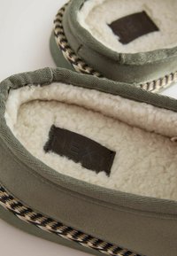 Roheline kangast suss, millel on pehme valge vooder, ja houndstooth aktsent talla ümber ning pruuni naha logo silt sees.