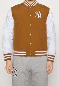 Brun och vit varsityjacka med randiga muddar och krage, knäppning med knappar och framträdande NY-logotyp på bröstet. Bärs med grå sweatshirtbyxor.