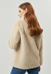 Beżowy płaszcz o teksturowanym wzorze, z pikowanym wzorem, miękkim materiałem i opadającymi ramionami. Stylizowany z niebieskimi dżinsami, widok od tyłu.