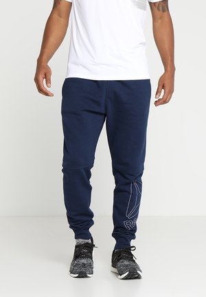 Man draagt marineblauwe joggingbroek met wit logo op linkerbeen, zwart-witte sneakers en een wit T-shirt, staat tegen een effen achtergrond.