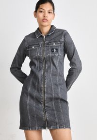 Denimkjole i mørkegrå, med glidelås foran, krage og to brystlommer. Den har vertikale sømdetaljer og en logo på lommen.