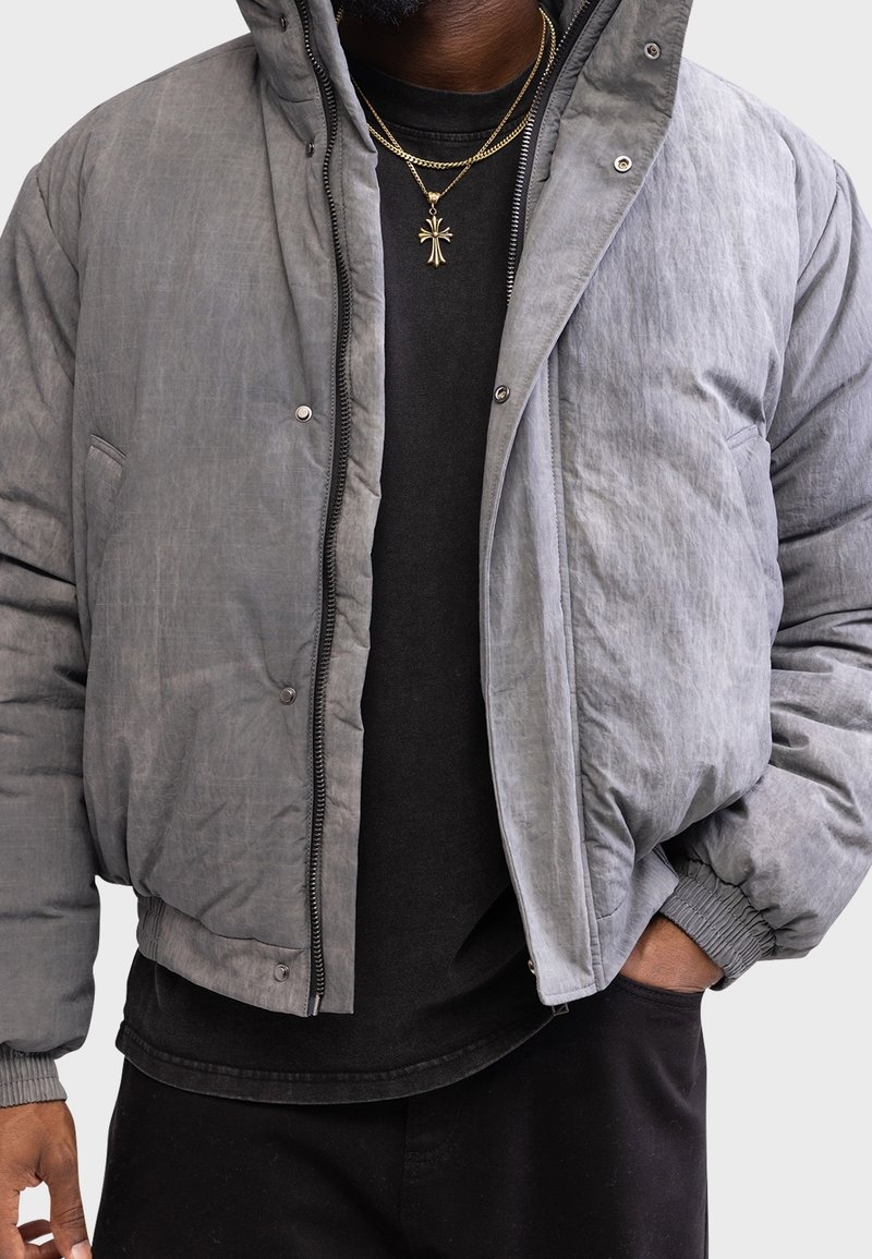 Grå pufferjakke med tekstureret finish, lynlåslukning, elastiske ærmekanter og lommer. Båret over en sort t-shirt med rund hals. Guld kædeaccenter synlige.