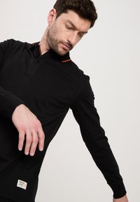 Czarna, długorękawowa koszulka polo z trzema guzikami, pomarańczowymi akcentami kołnierza oraz naszywką z logo u dołu. Gładki materiał bawełniany.