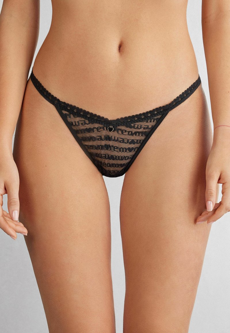 Zwarte kantstring met doorzichtig materiaal, golvende randen en een klein hartvormig bedeltje in het midden. Bevat een geometrisch patroon over de gehele lengte.