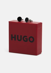Boutons de manchette Hugo en argent avec un cadran circulaire noir, arborant le logo "Hugo". Présentés dans une boîte rouge avec une écriture noire.