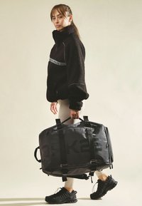 Nainen mustassa fleece-takissa ja vaaleissa legginsseissä kantaa suurta mustaa urheilukassia, jossa on "OAK25"-logo, jalassa mustat urheilukengät.