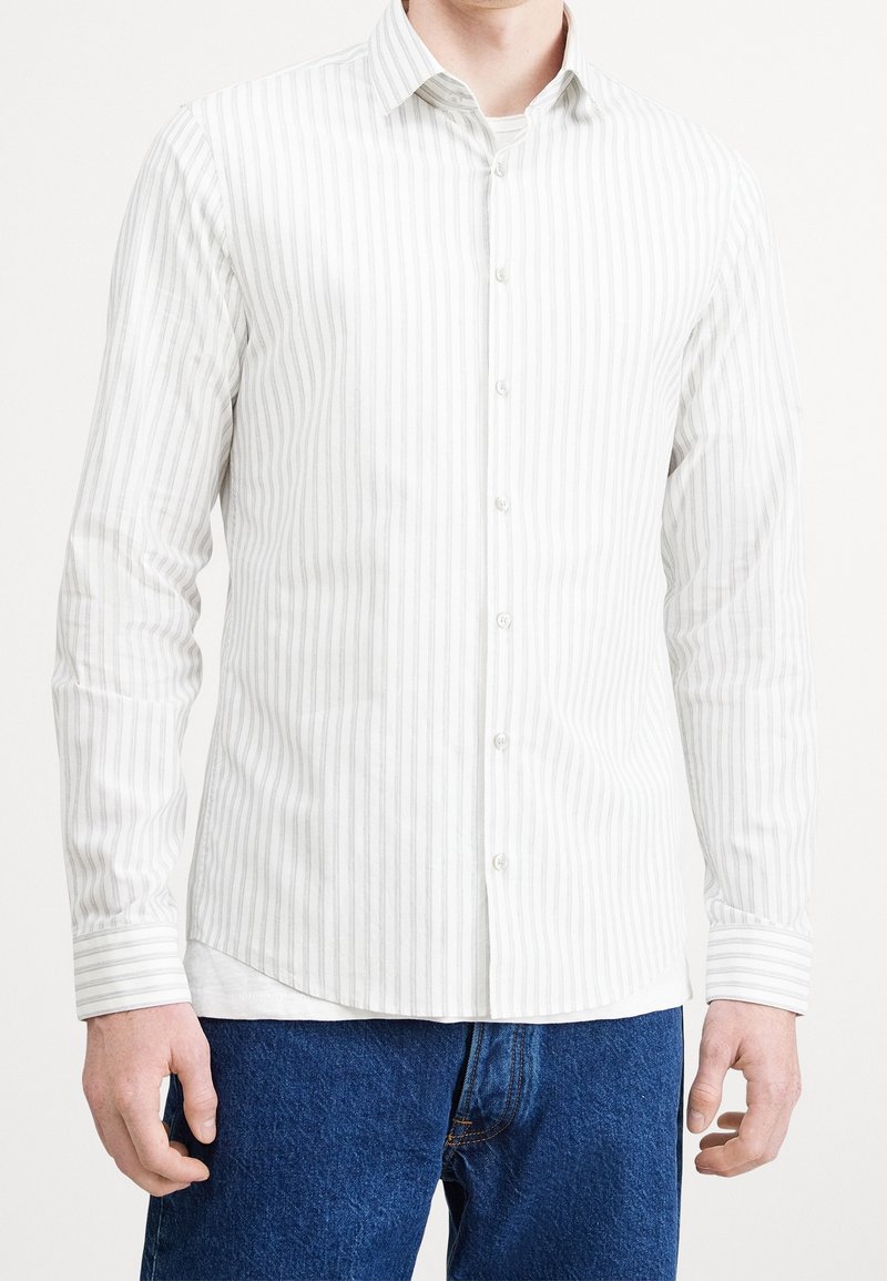 Bílá košile s dlouhým rukávem a šedými vertikálními pruhy, zapínání na knoflíky v přední části a klasický límec. Měkká tkanina s hladkým povrchem.
