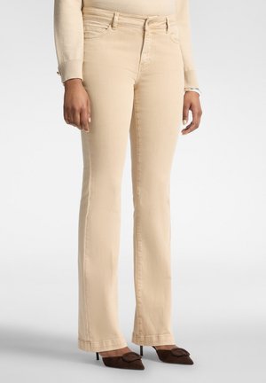 Donna che indossa pantaloni beige bootcut, top beige a maniche lunghe e tacchi scuri a punta, in piedi su uno sfondo neutro.