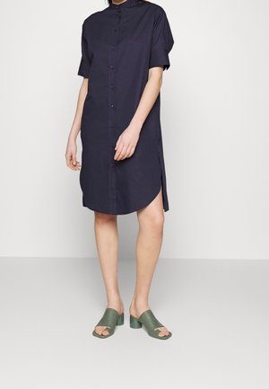 Person wearing a navy blue short-sleeve button-up shirt dress with side slits and green heeled open-toe sandals, standing on a plain background.