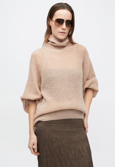 Világos bézs garbó laza szabással és bordázott mintával, sötétbarna pliszírozott szoknyával párosítva. Nő napszemüveget visel.