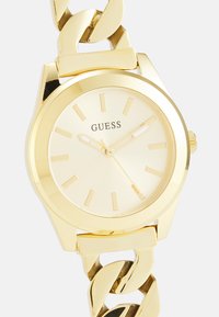 Gold-tone watch with large chain link strap. Round case, minimalistic dial with gold markers. Smooth, glossy finish, brand name displayed.