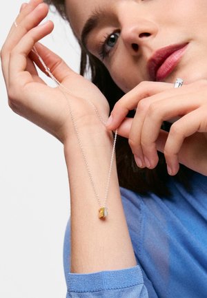 Persona con camisa azul sosteniendo un collar de cadena plateada con un pequeño colgante de mariposa dorada y plateada cerca de la muñeca.