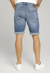 Pantalones cortos de denim en azul claro, con puños enrollados, dos bolsillos traseros y un parche de cuero marrón en la cinturilla. Llevados con zapatillas blancas.