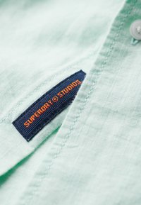 Light green fabric with close-up of a navy blue clothing label stitched on it, showing "SUPERDRY STUDIOS" in orange text.