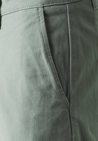 Tela de algodón verde con una textura suave, que presenta un bolsillo lateral y detalles de costura visibles. No hay patrones prominentes ni herrajes visibles.