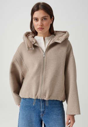 Jeune femme portant une veste beige zippée avec une capuche rembourrée et un jean bleu, debout devant un fond clair uni.