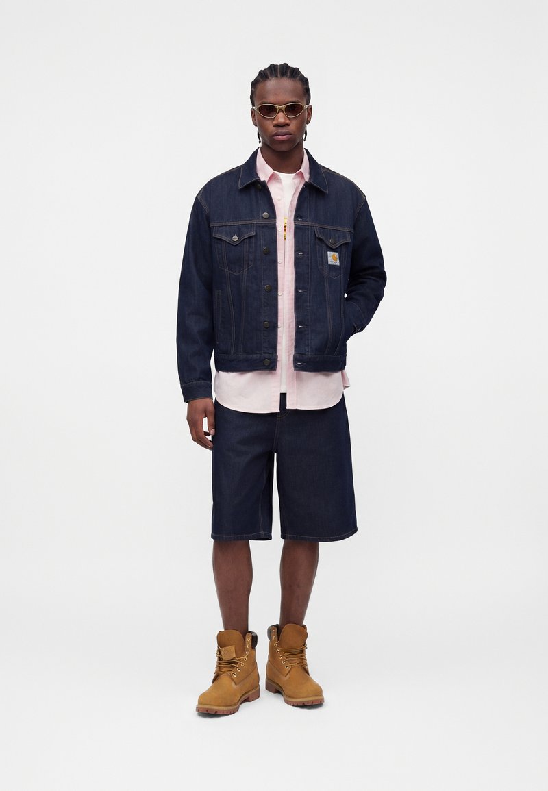 Man wearing sunglasses, dark denim jacket and shorts over light pink shirt with tan work boots, standing against plain background.