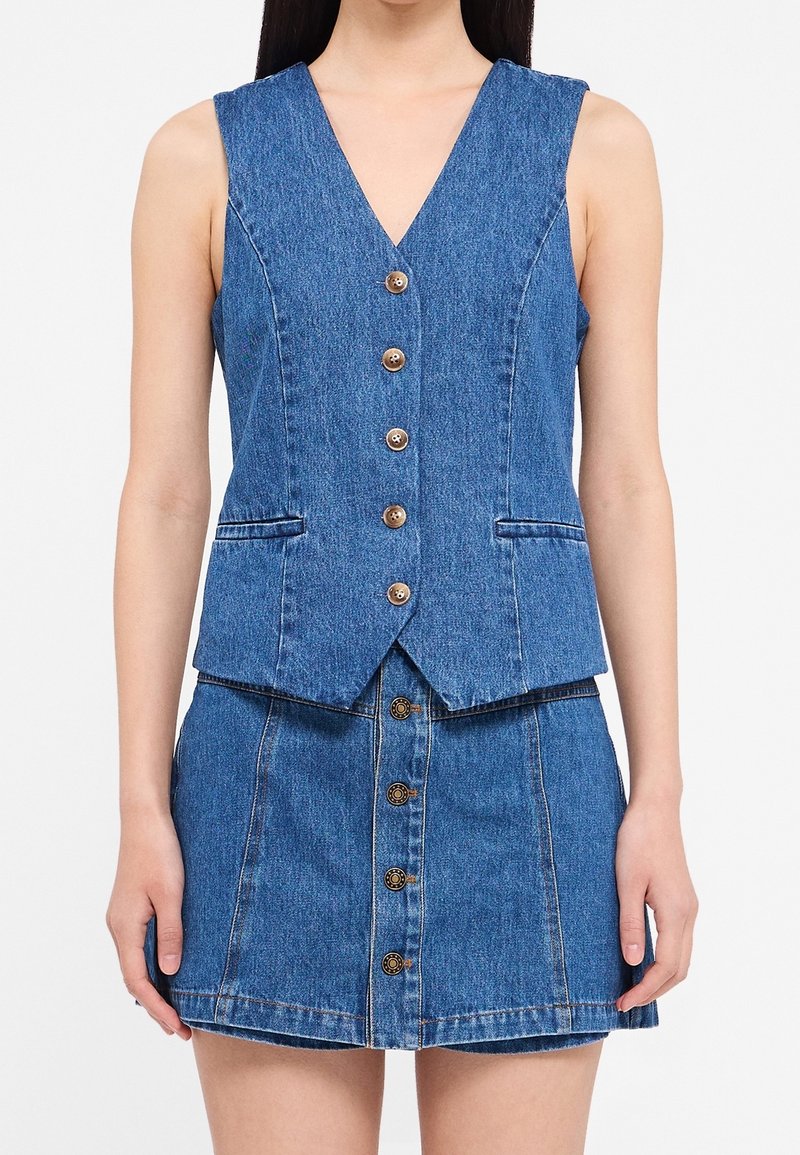 Denim-Weste und Rock-Set mit V-Ausschnitt, braunen Knöpfen und Kontrastnähten. Der Stoff ist blau, strukturiert und hat eine glatte Oberfläche.