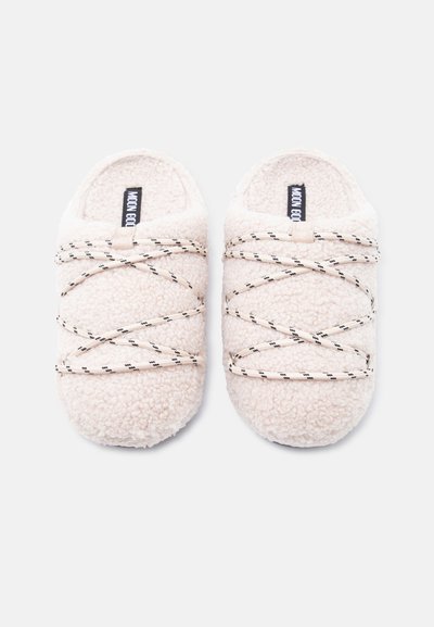 Chaussons beiges doux en tissu peluche, avec des lacets croisés noirs et beiges, un bout rond et un intérieur rembourré.