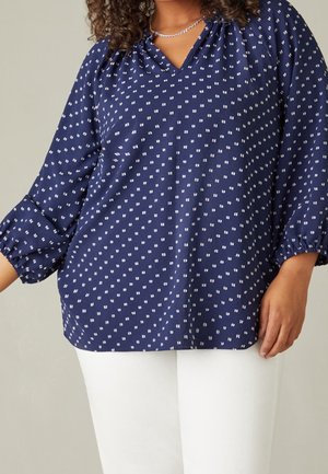 Nainen pukeutuneena laivastonsiniseen puseroon, jossa on valkoisia neliökuvioita, ja valkoisiin housuihin, seisomassa yksinkertaisen taustan edessä.