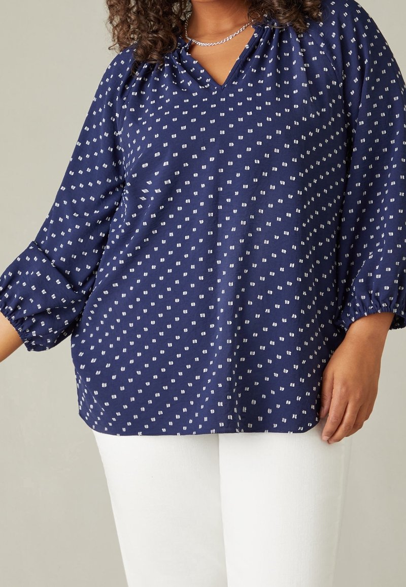 Frau trägt eine marineblaue Bluse mit weißen quadratischen Mustern und weiße Hosen, stehend vor einem schlichten Hintergrund.