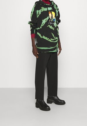 Personne portant un sweat à capuche noir tie-dye avec un motif Beavis et Butt-Head, un pantalon noir à jambes larges, des chaussures épaisses en cuir noir et un haut à manches longues rouge.