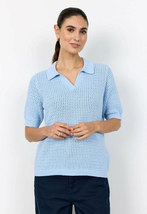 Vrouw met donker haar in een paardenstaart, gekleed in een lichtblauw gebreid poloshirt met korte mouwen en donkere broek, staand tegen een effen witte achtergrond.