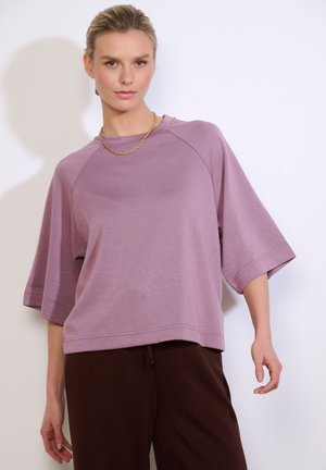 Woman wearing a loose mauve short-sleeve top, dark brown pants, and a gold chain necklace against a plain light background.