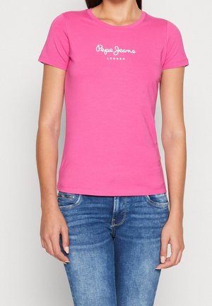 Kobieta ubrana w dopasowany różowy t-shirt z krótkim rękawem z logo "Pepe Jeans London" oraz niebieskie jeansy denim na jednolitym tle.