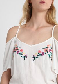 Bílá halenka s odhalenými rameny a krátkými rukávy. Na hrudi je zdobená barevným květinovým vyšíváním v růžové, žluté a fialové barvě. Texturovaná látka.