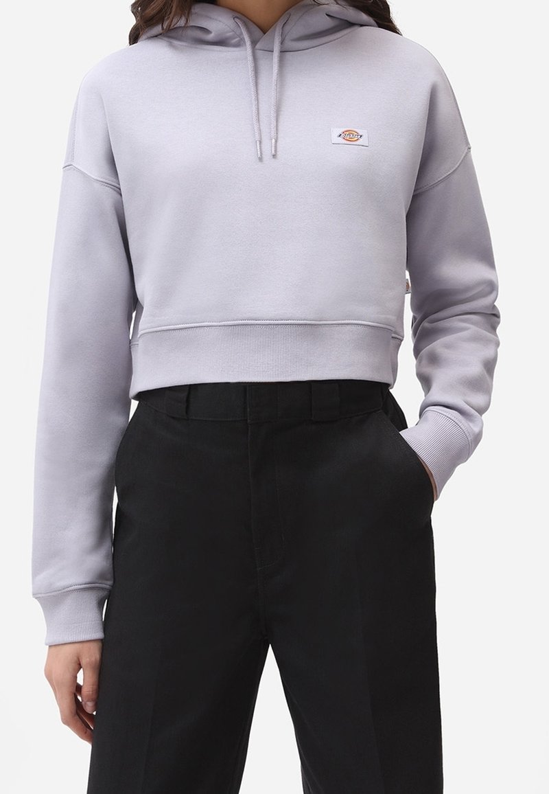 Personne portant un sweat à capuche court gris clair avec un petit logo sur la poitrine et un pantalon noir taille haute, une main dans la poche.