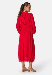 Rode jurk met lange mouwen en een strikdetail op de rug, met een smock taille. Gemaakt van een lichte stof, met een vloeiende lengte tot halverwege de kuit.