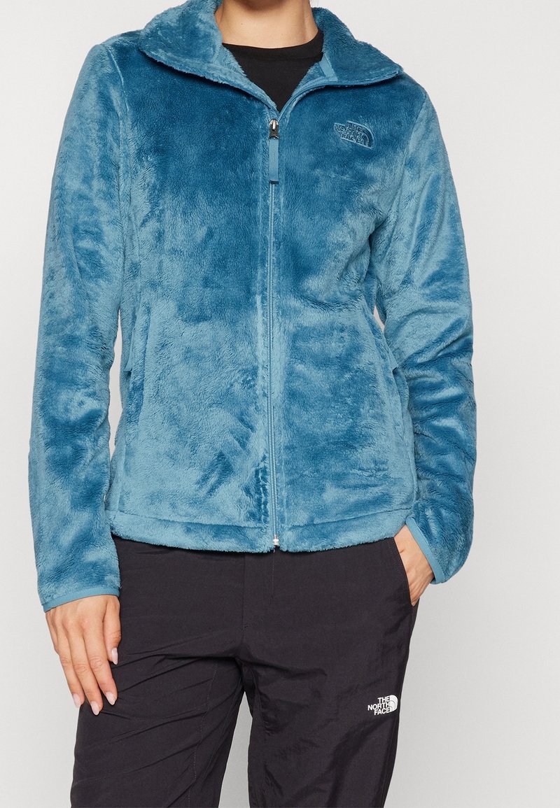 Personne portant une veste zippée en peluche bleue et un pantalon noir avec un logo sur les deux vêtements, debout avec une main dans la poche.