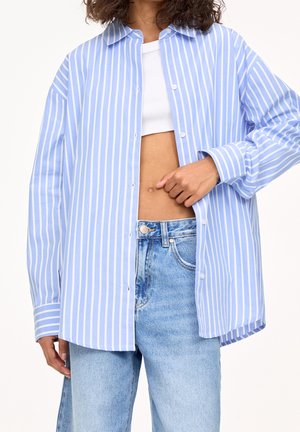 Person wearing loose blue and white vertical striped button-up shirt over white crop top and high-waisted light blue jeans, pointing at navel.