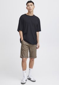 Camiseta negra de gran tamaño combinada con pantalones cortos beiges, puños enrollados, calcetines blancos de Nike y zapatillas grises/blancas de Adidas sobre un fondo sencillo.