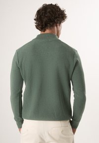 Grünes, geripptes Langarm-Polo mit Kragen, strukturiertem Stoff und figurnahem Schnitt, getragen zu hellen Hosen.