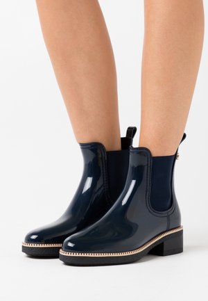 Bottines en vernis bleu marine avec des côtés élastiques, une semelle en caoutchouc noir texturé et un contour beige contrastant le long du bas.