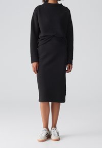 Pull en maille texturée noire associé à une jupe midi noire. Le modèle porte des baskets blanches avec des rayures noires et des semelles beiges.