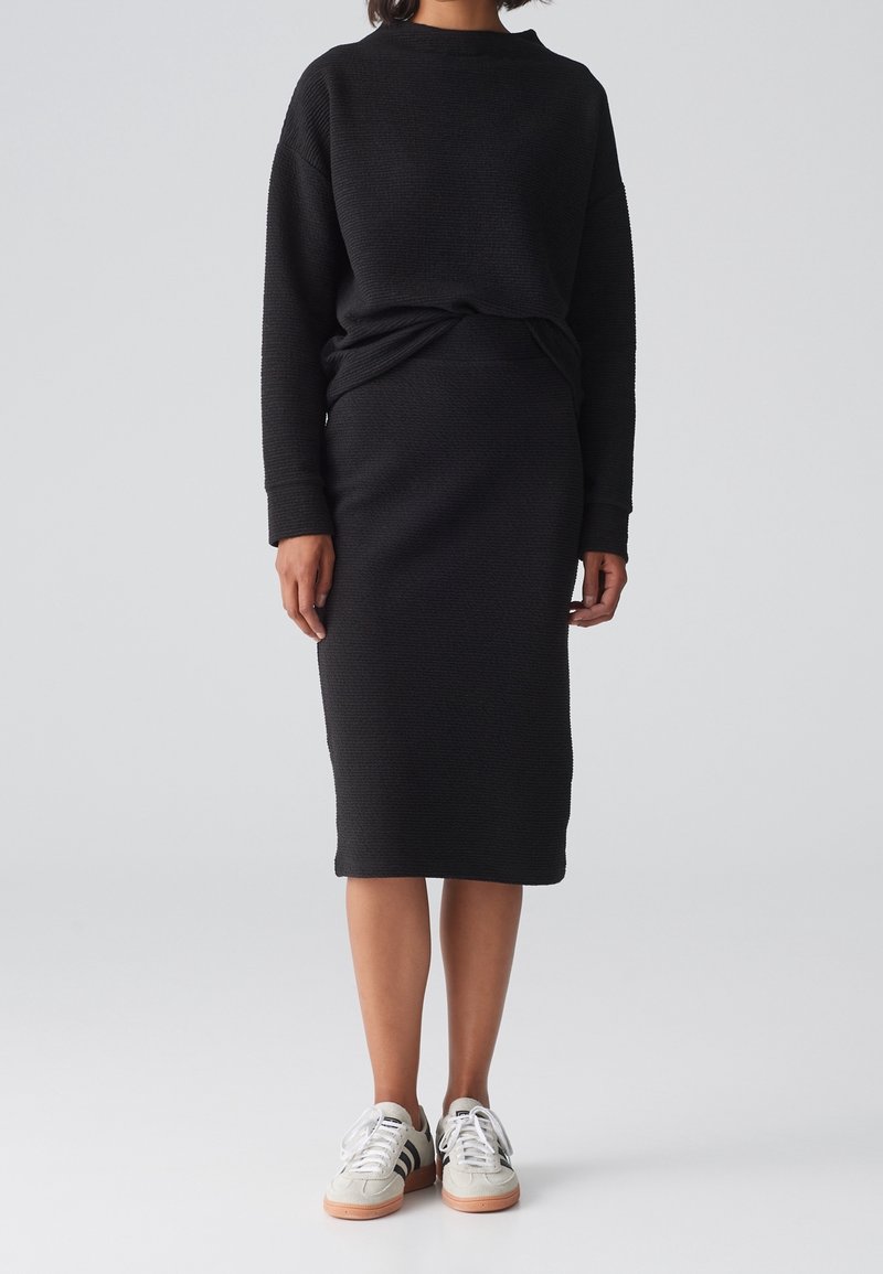 Pull en maille texturée noire associé à une jupe midi noire. Le modèle porte des baskets blanches avec des rayures noires et des semelles beiges.