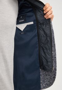 Giacca blu navy con esterno in tweed. All'interno presenta una fodera grigia, una tasca con zip e etichette, tra cui "JOOP!" e "SLIM FIT".