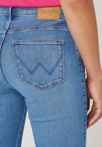 Vaqueros de mezclilla azul claro con corte recto, que presentan un distintivo patrón de costura en forma de "W" en el bolsillo trasero y una etiqueta de cuero marrón.