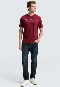 Maroon cotton t-shirt with white printed text, paired with dark blue denim jeans and white sneakers. Model stands casually with one hand in pocket.